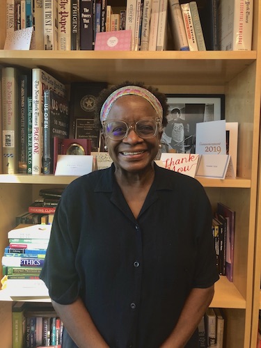 Portrait photo of director of the School of Journalism and Media, Kathleen McElroy, standing in front of the bookshelf in her office. She is a Black woman and is wearing a Black blouse, tan glasses and a headband. She stands smiling with her hands crossed in front of her. 