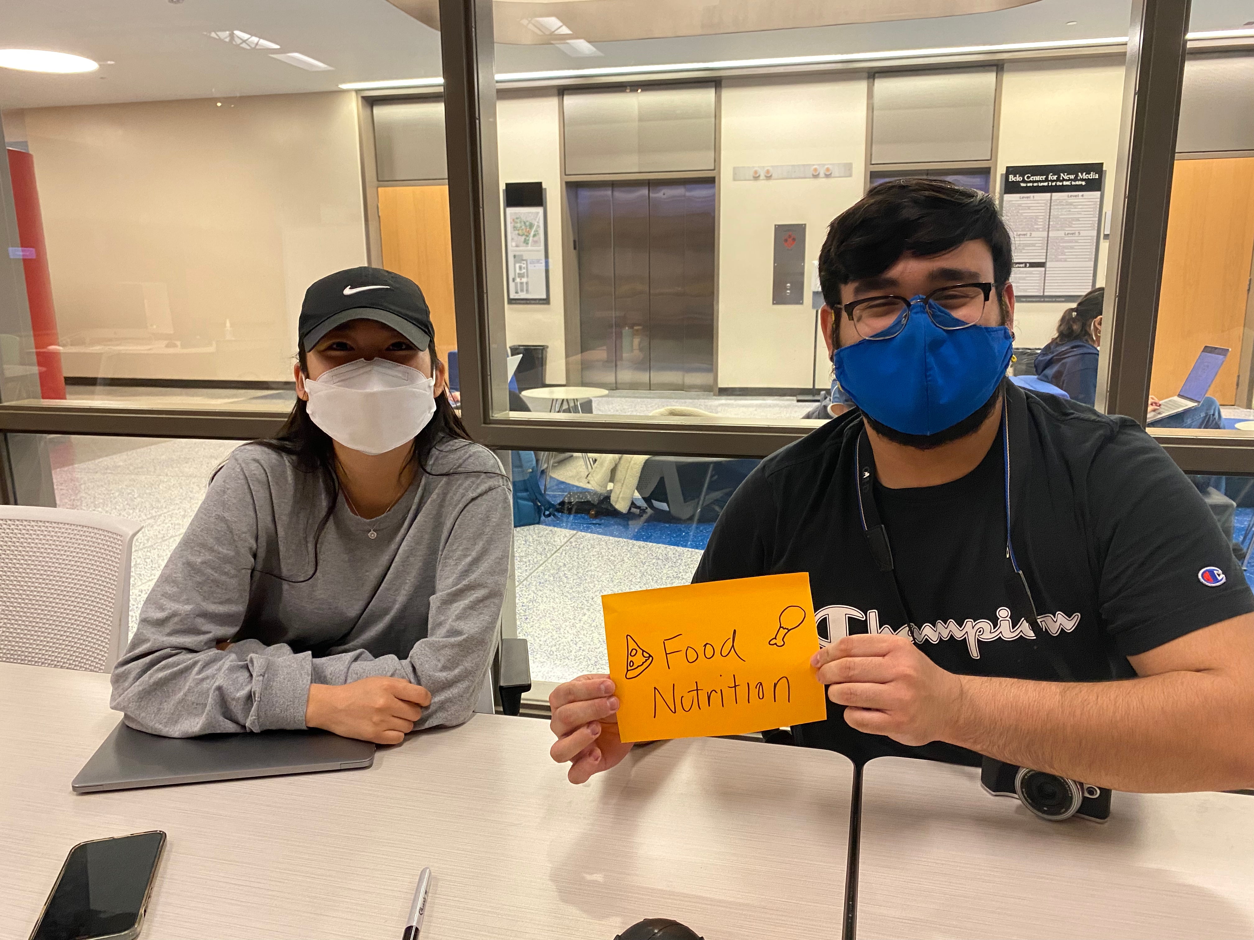 Two students sit at a desk holding a sign that says Food and Nutrition