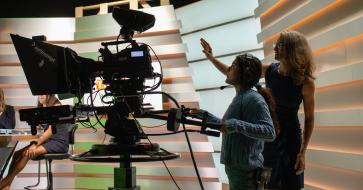 A student stands behind a broadcast camera and is being directed by Professor Kate West.