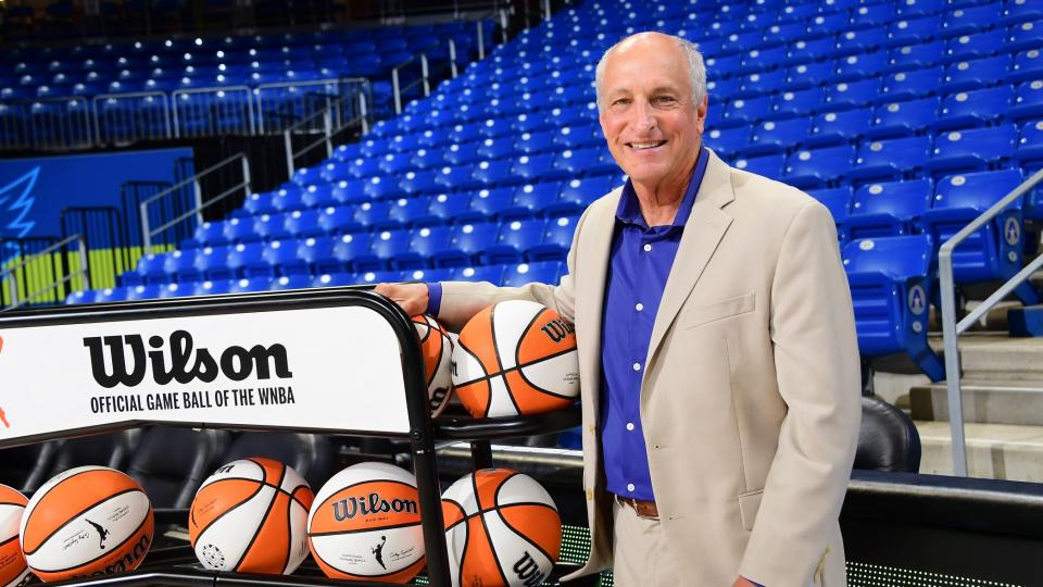 Robert Steinfeld stands next to a rack of Wilson basketballs in an arena with blue stadium seats.