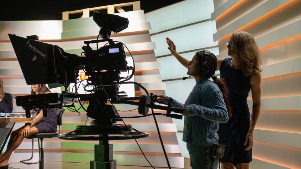 A student stands behind a broadcast camera and is being directed by Professor Kate West.