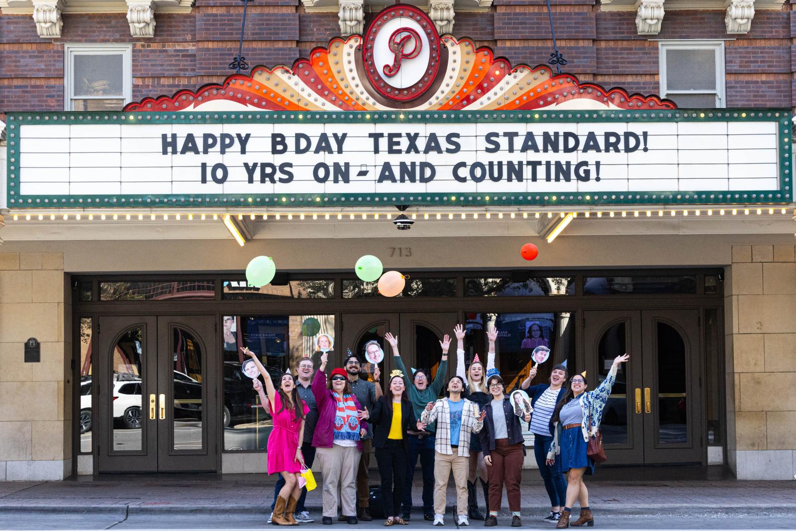 A group of people stands under the marquee at the Paramount Theater with their arms raised in celebration. The marquee reads "Happy bday Texas Standard! 10 yrs on - and counting!"
