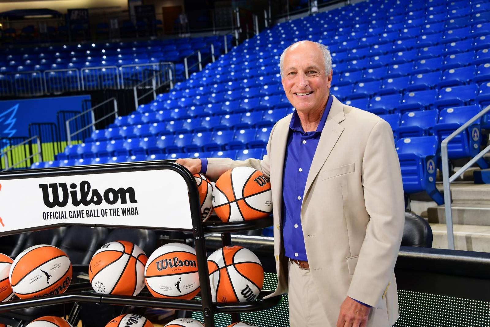 Robert Steinfeld stands next to a rack of Wilson basketballs in an arena with blue stadium seats.