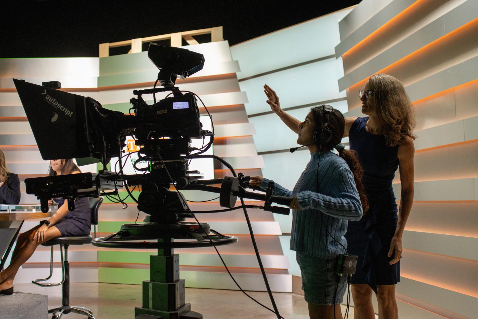 A student stands behind a broadcast camera and is being directed by Professor Kate West.