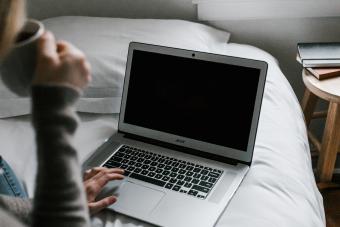 A woman sips coffee while sitting on a bed with an open laptop